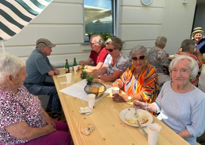 Seniorinnen und Senioren sitzen an einem langen Holztisch im Freien beim Grillfest; im Vordergrund rechts eine Frau isst, mittig eine Frau in buntem Kleid mit Sonnenbrille.