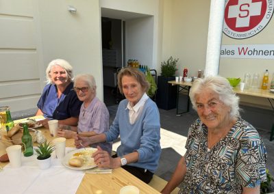 Vier Damen sitzen an einem Holztisch und essen; Getränke und Samariterbund-Logo im Hintergrund.
