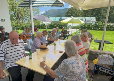 Ältere Gäste an einem langen Tisch unter Sonnenschirmen, plaudern und trinken; Musiker mit Ziehharmonika und Gitarre im Hintergrund; Wandtext ‚30A‘ sichtbar.