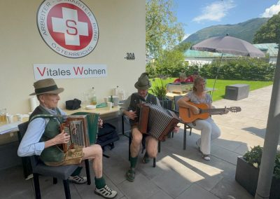 Drei Musiker auf einer Terrasse, zwei mit Ziehharmonika, eine mit Gitarre; an der Wand Samariterbund-Logo und Schild "Vitales Wohnen"; Gäste, Wiese und Bergkulisse im Hintergrund.