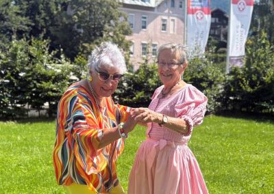 Zwei ältere Frauen tanzen lächelnd auf einer sonnigen Wiese; im Hintergrund sind Rotkreuz-Fahnen sichtbar.