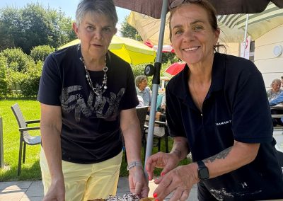 Zwei Frauen unter Sonnenschirmen richten Kuchenstücke auf einer Servierplatte an; auf dem Polo einer Frau ist „Samarit“ sichtbar; Gäste im Hintergrund.