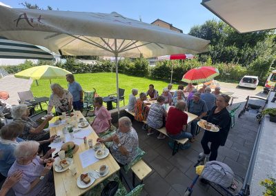 Ältere Menschen sitzen an zwei langen Holztischen im Garten unter bunten Sonnenschirmen; Tassen, Kuchen und Teller auf den Tischen, eine Mitarbeiterin trägt ein Tablett.