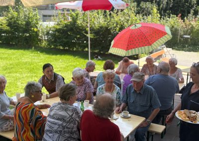Ältere Menschen sitzen an Holztischen im Garten unter bunten Sonnenschirmen; eine Mitarbeiterin serviert Kuchen.