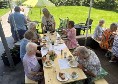 Seniorinnen und Senioren an langen Holztischen im Garten, trinken Kaffee und essen Kuchen; Sonnenschirme und Grill im Hintergrund.