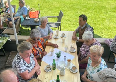 Mehrere ältere Personen sitzen an einem langen Holztisch auf einer Terrasse; auf dem Tisch Kaffeetassen, Pappbecher, Flaschen und ein Stück Kuchen; Rasen im Hintergrund.