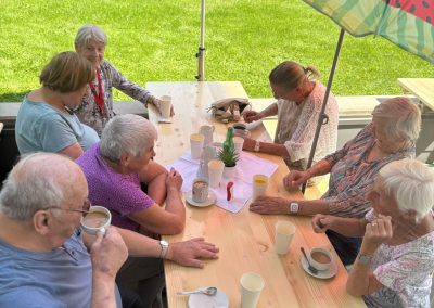 Sieben ältere Erwachsene sitzen an einem langen Holztisch im Freien unter einem bunten Sonnenschirm (Wassermelonenmuster); auf dem Tisch Tassen, Pappbecher, Teller und eine kleine Pflanze; im Hintergrund Rasen.