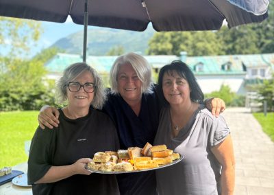 Drei Frauen unter einem Sonnenschirm halten ein Tablett mit Kuchenstücken und lächeln.
