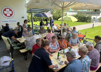 Mehrere ältere Menschen sitzen draußen an langen Tischen unter Sonnenschirmen, trinken Kaffee und essen Kuchen; an der Wand sind Schriftzug „Vitales Wohnen“ und ein Arbeiter‑Samariter‑Bund‑Logo sichtbar.