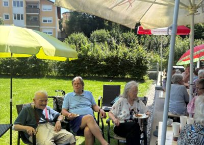 Teilnehmende sitzen unter Sonnenschirmen im Garten und essen Kuchen; auf einem Schirm ist der Schriftzug ‚Art‘ sichtbar.