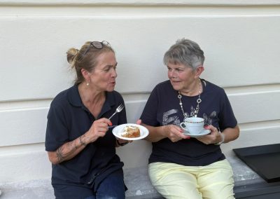 Zwei Frauen sitzen auf einer niedrigen Mauer vor heller Fassadenwand; links Pappteller mit Kuchen und Gabel, rechts Tasse mit Untertasse.