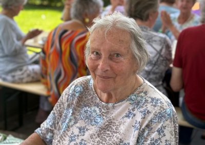Ältere Frau sitzt draußen an einem Tisch mit Kaffee und Kuchen, im Hintergrund Gäste bei einem Fest