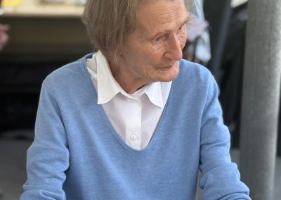 Ältere Frau in hellblauem Pullover sitzt am Tisch, schaut nach rechts; Tasse vor ihr.