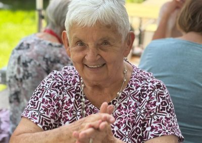 Lachende Teilnehmerin beim Grillfest, sitzt an einem Tisch im Freien, Hände vor dem Körper sichtbar.