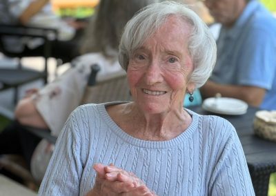 Ältere Frau sitzt lächelnd bei einem Außentreffen, weitere Personen unscharf im Hintergrund.