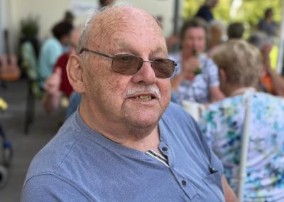 Älterer Mann mit Sonnenbrille sitzt an einem Tisch und lächelt; unscharfer Hintergrund mit weiteren Gästen und einem runden Schild mit rotem Kreuz.
