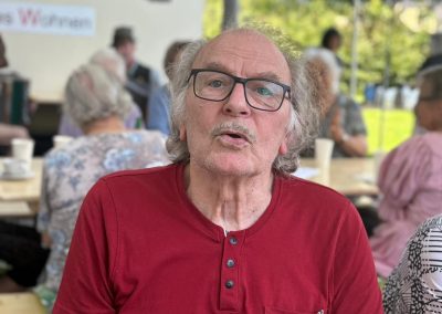 Ein Mann mit Brille in rotem T-Shirt sitzt an einem Festtisch; im Hintergrund Gäste und ein Rotkreuz‑Logo.