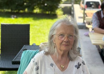 Frau sitzt im Freien auf einem Stuhl, trägt Brille und ein T‑Shirt mit großem Katzenmotiv.