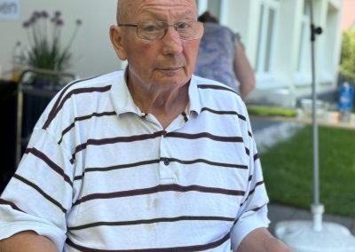 Älterer Mann sitzt im Schatten vor einem Gebäude, trägt gestreiftes Poloshirt und Brille.