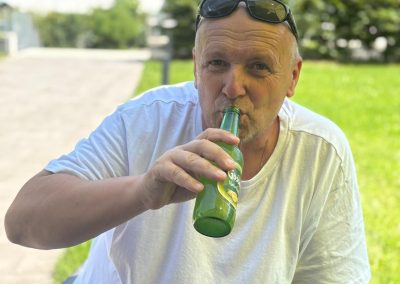 Mann in weißem T‑Shirt sitzt draußen, trinkt aus einer grünen Flasche; Sonnenbrille auf dem Kopf, Flaschenetikett mit Zitronenmotiv sichtbar.