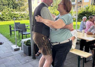 Zwei Personen tanzen beim Outdoor-Grillfest: Mann in Lederhose und Hut, Frau im T‑Shirt; im Hintergrund Sitzgruppen und Sonnenschirm.