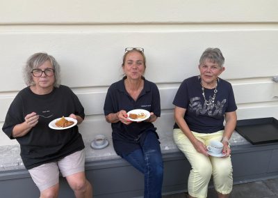 Drei Frauen sitzen nebeneinander an einer hellen Wand, essen Kuchen von Papptellern und trinken Kaffee; die mittlere trägt ein T‑Shirt mit kleinem Schriftzug.