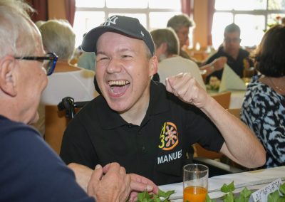Lachender Mann mit Baseballcap und schwarzem Polo mit Aufschrift "MANUEL", sitzt an gedecktem Tisch und gestikuliert.