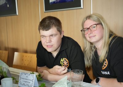 Zwei Personen (junger Mann, junge Frau mit Brille) an gedecktem Tisch, schwarze Poloshirts, Tischkarte mit Text 'M3 Mitarbeiter/innen + Angehörige'.