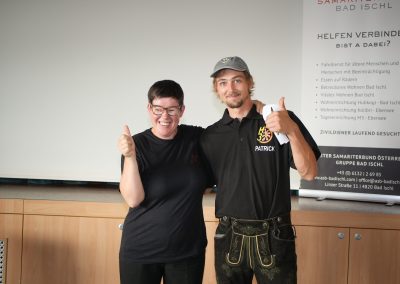 Zwei lächelnde Personen stehen nebeneinander und zeigen Daumen hoch; rechts schwarzes Polo mit weißem Schriftzug sichtbar.