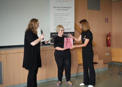Drei Frauen auf einer Bühne; links eine Frau mit Mikrofon und Zettel, in der Mitte eine Frau, die eine rosa Muster-Geschenktüte entgegennimmt, rechts eine Frau, die die Tüte überreicht.