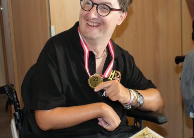 Person im Rollstuhl, lächelnd, hält Medaille mit Aufschrift '23 Jahre' und eine Urkunde.