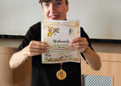 Mann in schwarzem T‑Shirt und Cap hält eine Urkunde mit dem Titel „Urkunde“ und eine Medaille mit der Aufschrift „25 Jahre“.