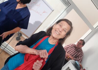 Lächelnde ältere Frau hält rotes Tuch; im Hintergrund zwei Personen und ein Flipchart mit teilweise lesbarem "Willkommen".