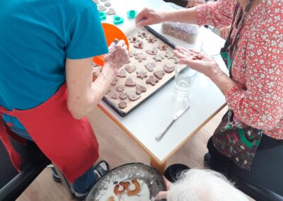 Mehrere ältere Personen an einem Tisch beim Ausstechen und Bestäuben von Plätzchen; Backblech mit ausgestochenen Keksen und eine Schüssel mit Puderzucker.