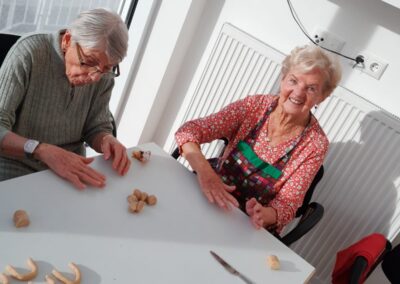 Zwei ältere Frauen sitzen an einem Tisch und formen kleine Teigstücke zu Hörnchen; die rechte Frau lächelt, die linke trägt eine Brille.
