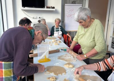 Ältere Frauen an einem langen Tisch rollen und stechen Teig zu Keksen; auf einem Flipchart steht handschriftlich ‚Herzlich Willkommen‘.