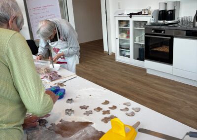 Zwei Personen an einem Küchentisch rollen Teig aus und stechen Plätzchen aus; bemehlter Tisch, Ausstechformen, Messbecher; Backofen und Flipchart im Hintergrund.