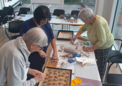 Drei Personen bereiten Plätzchen an einem Tisch vor; Backbleche, Ausstechformen, Nüsse und eine Schüssel mit Ei-Wasch sichtbar.