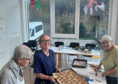 Drei Frauen in einem hellen Raum bereiten Gebäck vor; die Frau in der Bildmitte hält ein Backblech mit ungebackenen Keksen, im Hintergrund große Fenster und ein geparkter Transporter.