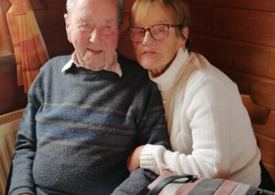 Zwei Personen sitzen nebeneinander an einem gedeckten Tisch in einem holzvertäfelten Raum; auf dem Tisch Handtasche, Besteck und grüne Glasflaschen.