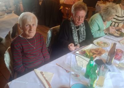 Zwei ältere Frauen an einem gedeckten Tisch in einem Restaurant, im Vordergrund Teller mit Schnitzel, Salat und Getränken; eine Frau blickt in die Kamera.