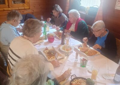 Sechs ältere Personen sitzen an einem gedeckten Tisch in einem holzverkleideten Raum und essen; auf dem Tisch Teller mit Pommes, einem Pfannengericht und Getränken.
