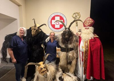 Zwei Personen in blauer Arbeitskleidung neben drei Krampus-Kostümen und einer Person in roter Bischofsrobe mit Stab; an der Wand das Logo 'Arbeiter-Samariter-Bund Österreichs'.