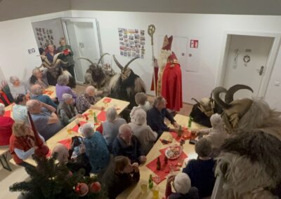 Gemeinschaftsraum: Nikolaus in rotem Gewand mit Bischofsstab und mehrere Krampus-Figuren; Seniorinnen und Senioren sitzen an Tischen mit Getränken und Snacks. Sichtbarer Text: 'EG'.