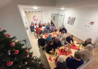 Vogelperspektive auf mehrere ältere Menschen an zwei langen Tischen mit roten Servietten und Getränken; links ein geschmückter Weihnachtsbaum; im Hintergrund Banner mit rotem Logo und Text.