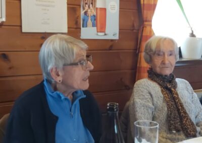 Zwei ältere Frauen sitzen an einem gedeckten Tisch in einem holzverkleideten Innenraum; im Vordergrund Bierflasche und -glas; Poster mit Text ‚DEIN BIER, DEIN REVIER!‘ im Hintergrund.