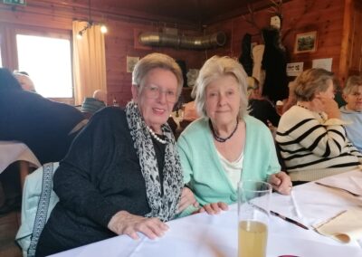 Zwei ältere Frauen sitzen nebeneinander an einem gedeckten Tisch in einem holzvertäfelten Gastraum; grünes Notausgangszeichen sichtbar.