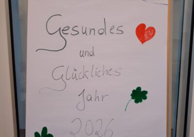 Flipchart mit handschriftlicher Botschaft 'Gesundes und Glückliches Jahr 2026', zwei rote Herzen, zwei grüne Kleeblätter; Partyhüte mit Aufschrift 'Happy New Year' oben.