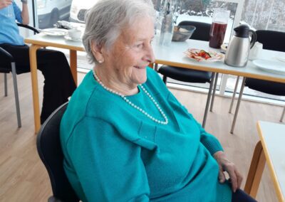 Ältere Frau in türkisfarbenem Pullover mit Perlenkette sitzt seitlich in einem Stuhl, lächelnd; im Hintergrund Tische mit Kaffeekanne, Snacks und großen Fenstern mit verschneiter Außenansicht.