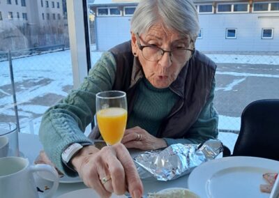 Person mit Brille greift am gedeckten Frühstückstisch mit einem Messer in einen Aufstrichbecher; im Hintergrund Fenster mit verschneiter Außenansicht und Gebäuden.